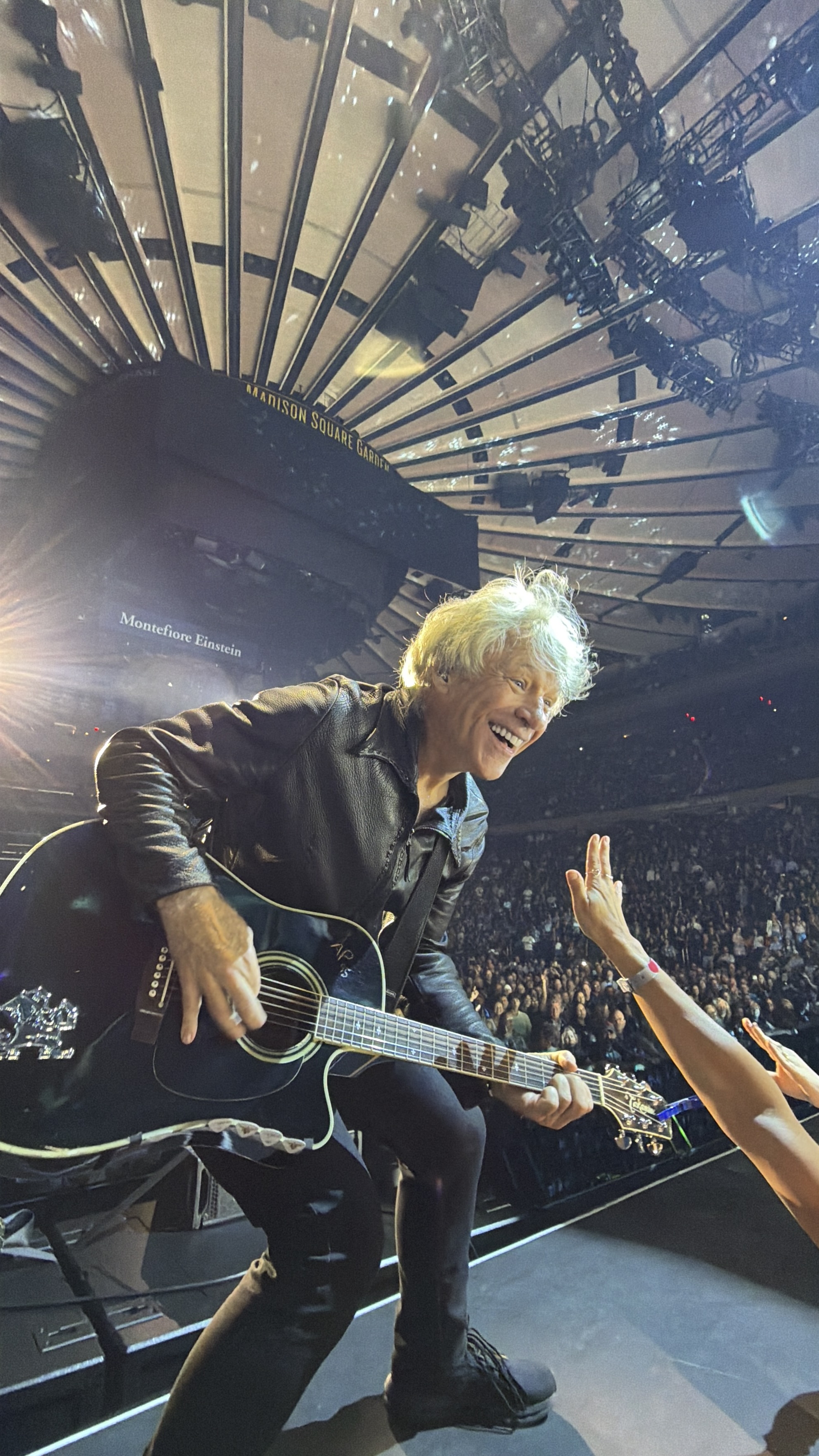 Jon Bon Jovi no palco, guitarra na mão, cabelos grisalhos, capturado por uma câmera de celular na plateia.